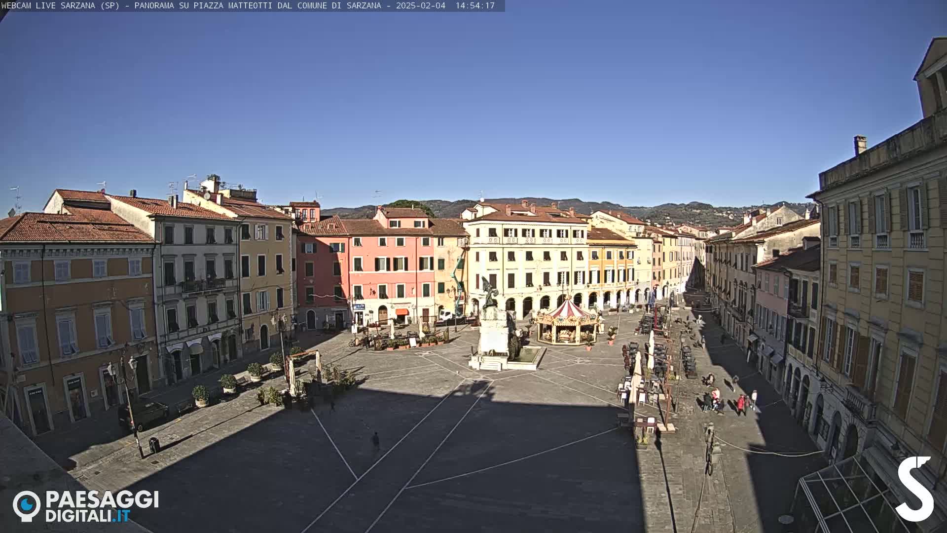 A sunny day reveals a large town square surrounded by multi-story buildings, a central monument, and a small carousel.