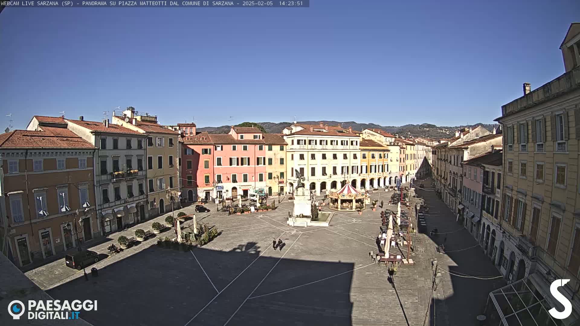A mostly sunny day reveals a large, empty town square surrounded by multi-story buildings, featuring a central monument and a carousel.
