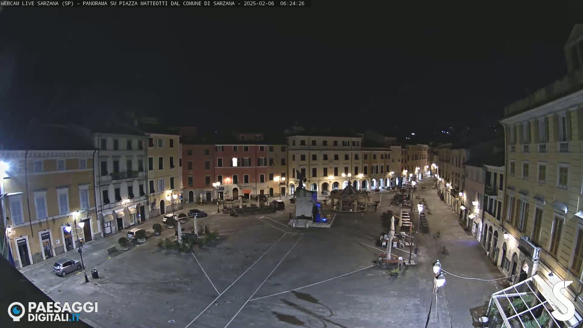 A nighttime, high-angle view of a sparsely populated town square, featuring several buildings with illuminated windows and a central fountain.