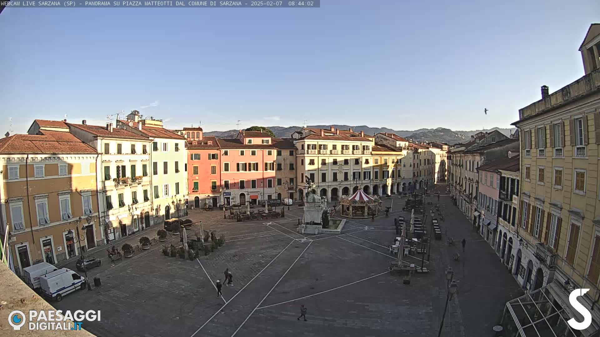 A mostly empty town square, surrounded by pastel-colored buildings under a clear blue sky, is shown in a high-angle, long shot.