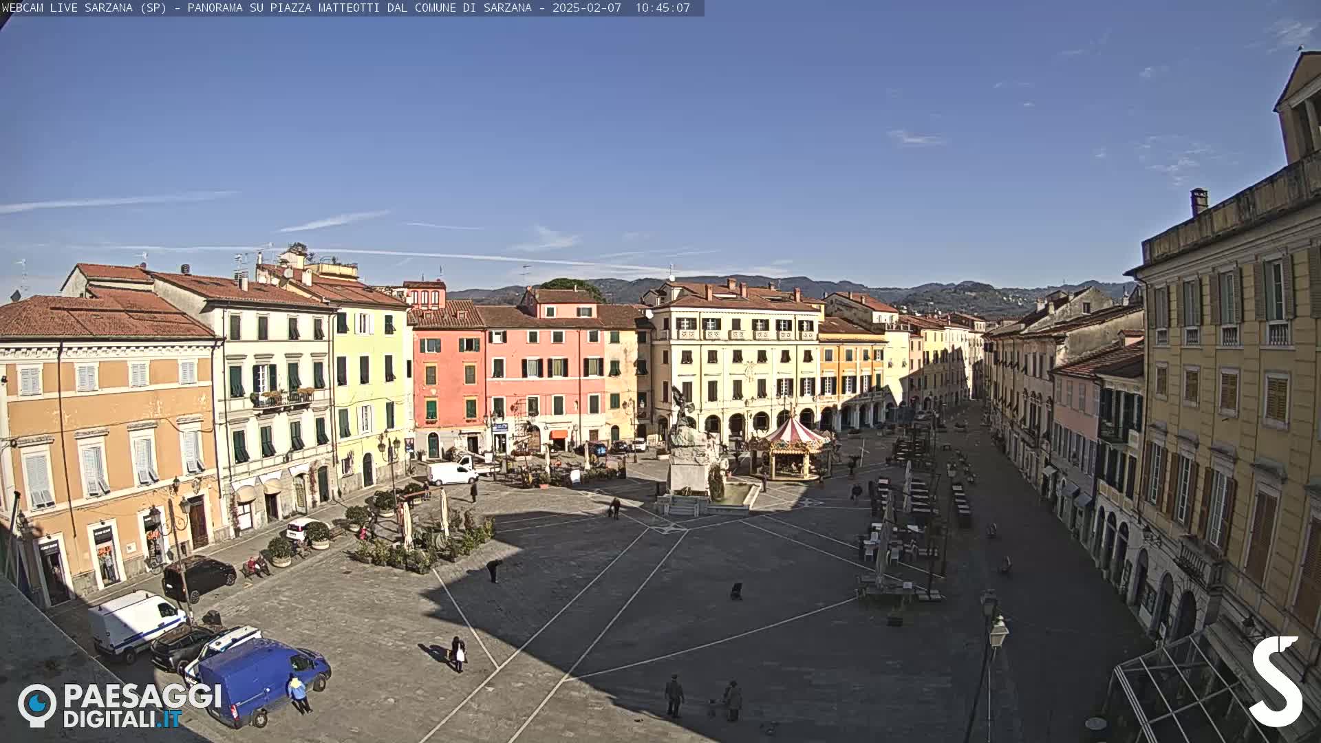 A large town square, surrounded by multi-story buildings under a clear blue sky, is sparsely populated with a few people and vehicles.