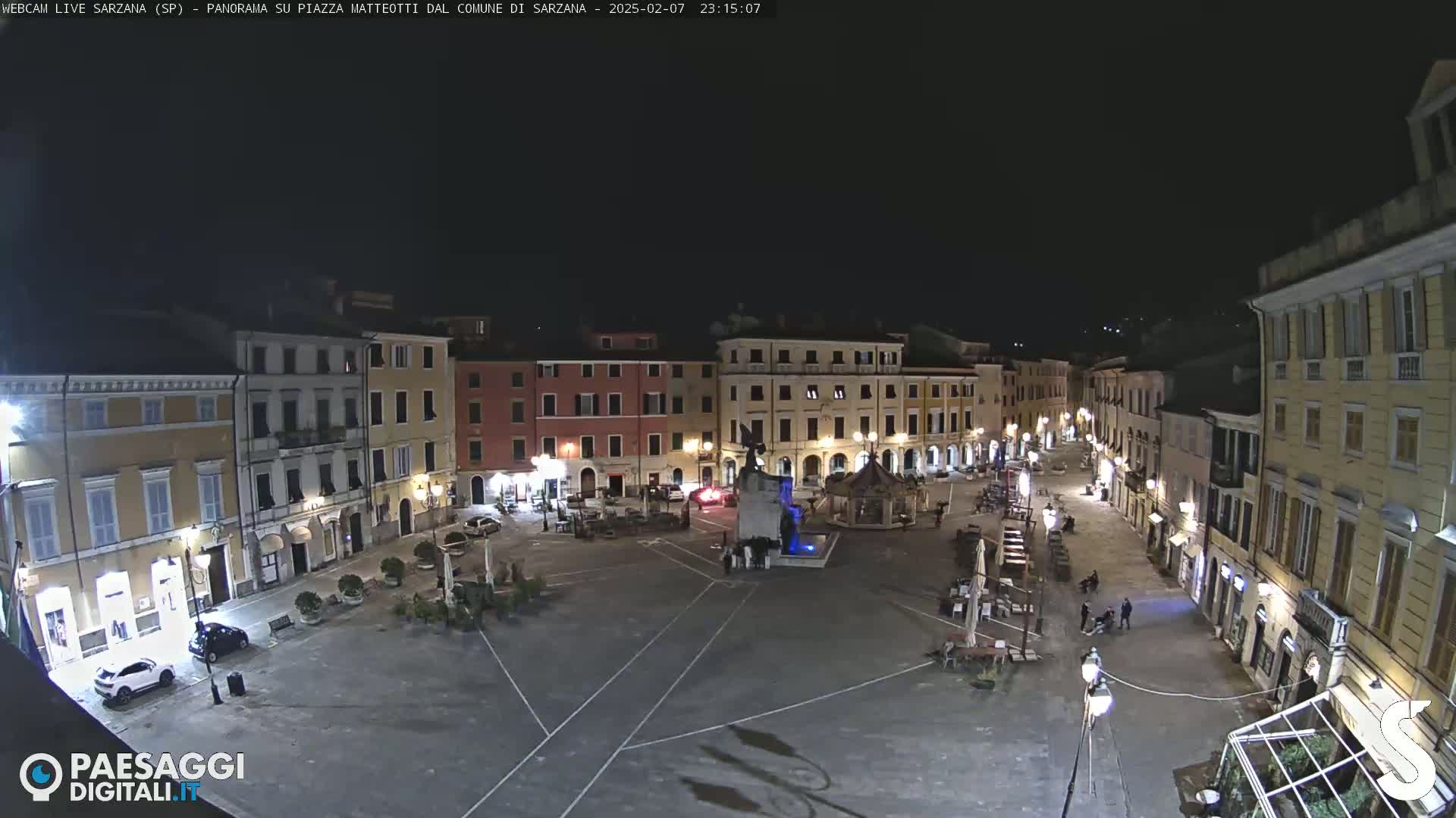 A nighttime, mostly empty town square is surrounded by multi-story buildings with lights illuminating the area.