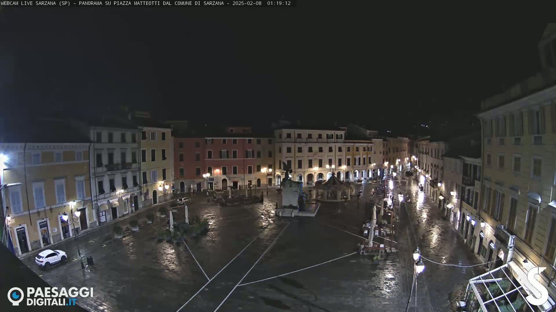 A rain-slicked, nighttime view of a town square surrounded by buildings, featuring a monument in the center.