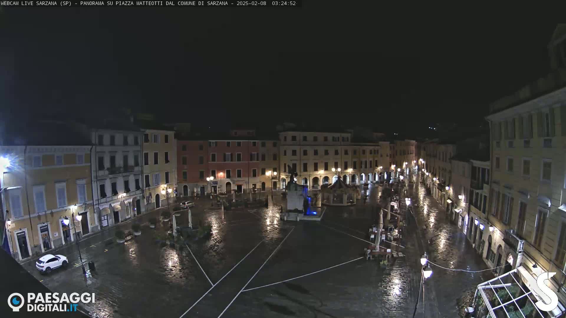 A rain-slicked town square at night, surrounded by illuminated buildings.
