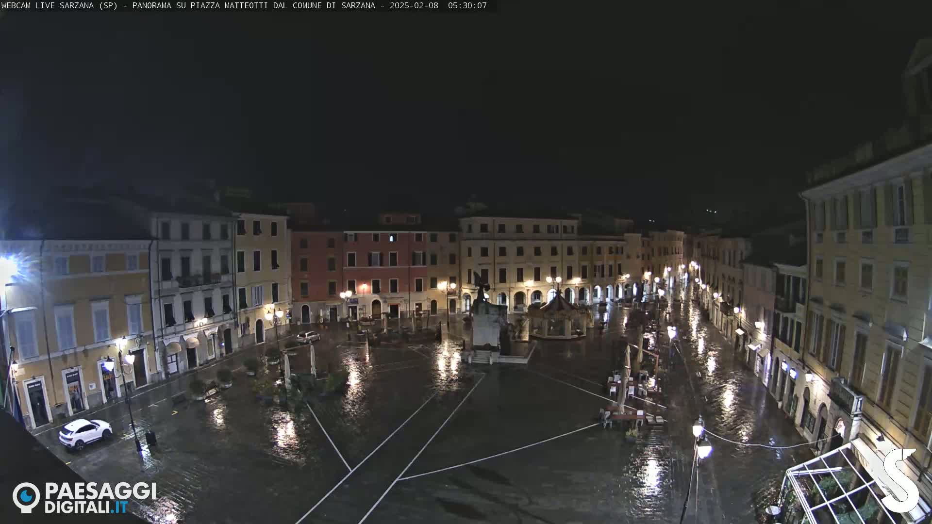 A floodlit town square, submerged in rainwater at night, is surrounded by buildings.
