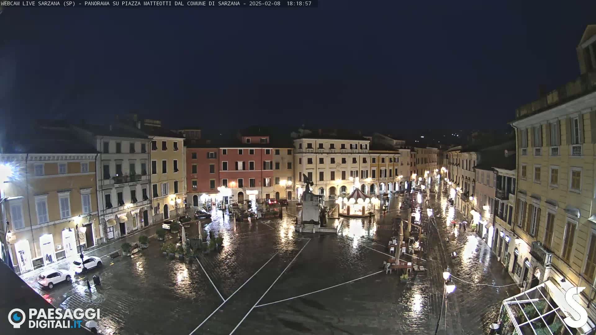 A rain-slicked town square at night, surrounded by illuminated buildings, features a central monument and a few cars parked near the edges.