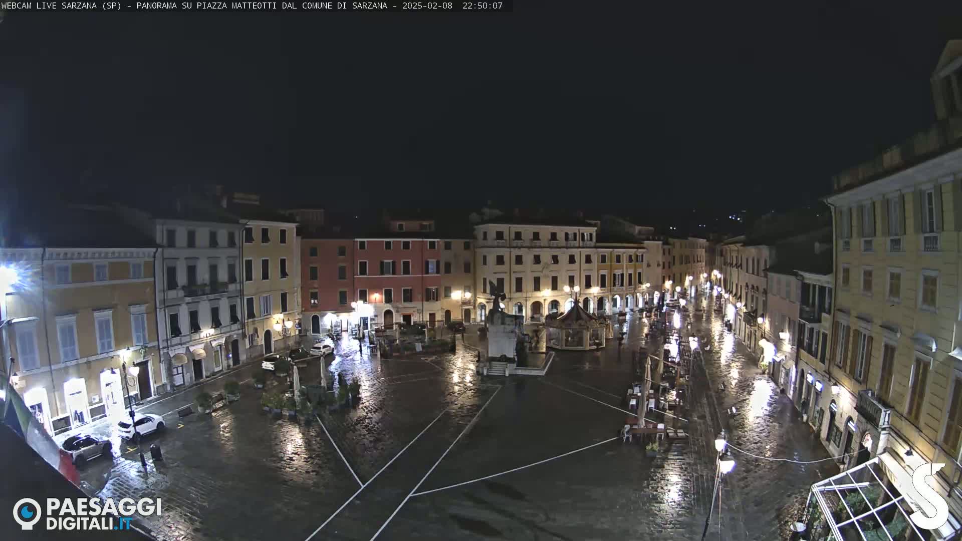 A rain-slicked town square at night, surrounded by illuminated buildings.