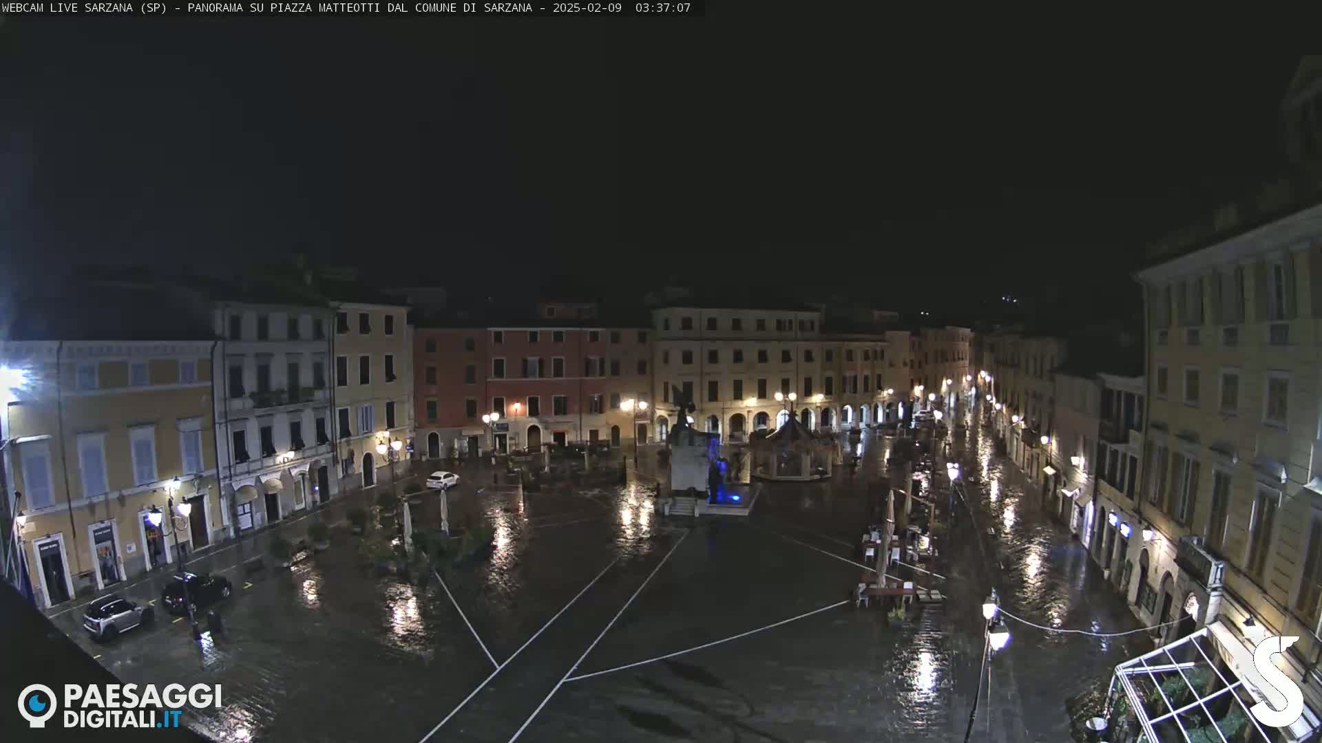A nighttime view of a flooded town square surrounded by buildings under heavy rain.