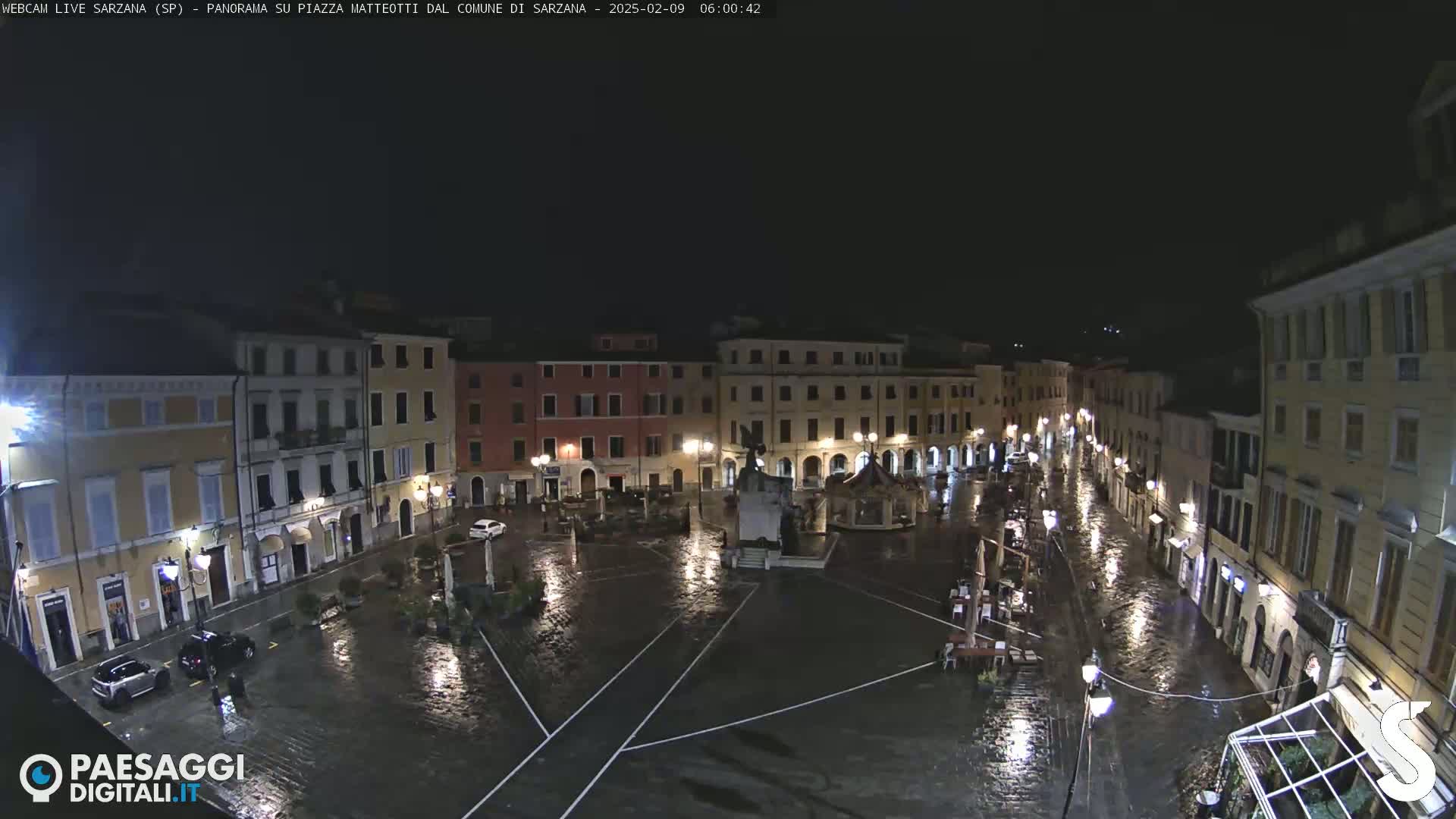 A rain-slicked town square at night, surrounded by illuminated buildings, with a few cars parked and some outdoor furniture visible.