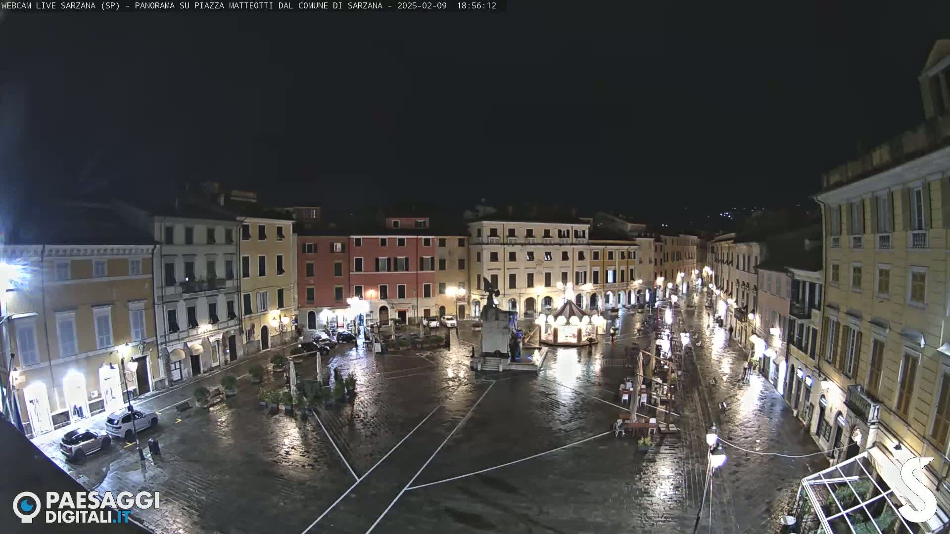 A rain-slicked, nighttime town square is surrounded by multi-story buildings with illuminated windows and streetlights.