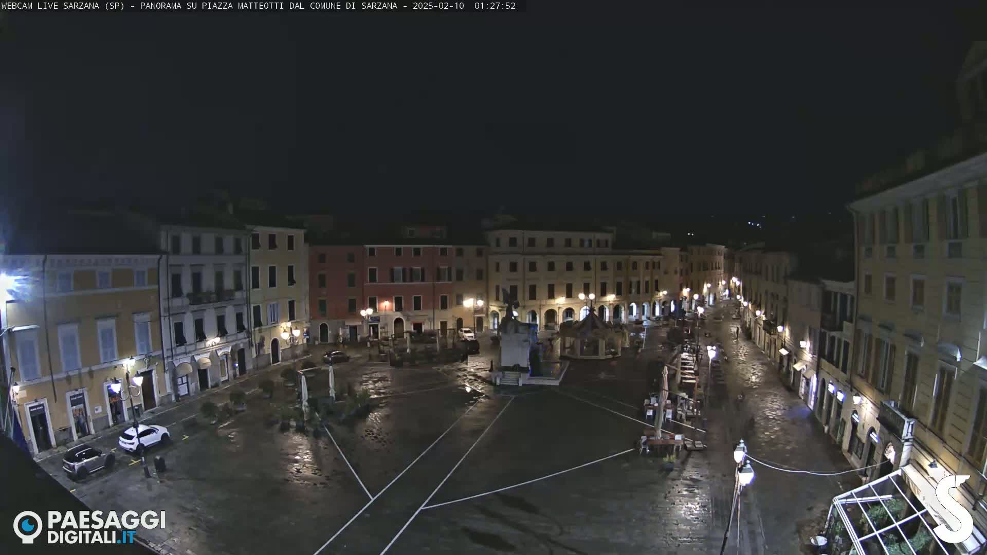 A wet, empty town square at night, surrounded by buildings with illuminated windows.