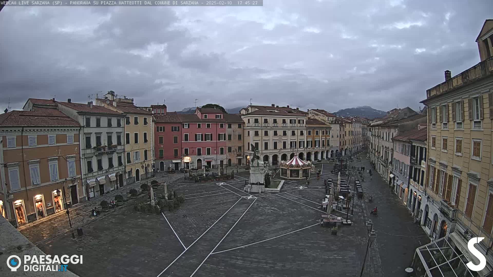 A mostly empty town square with multi-story buildings surrounding it under an overcast sky.