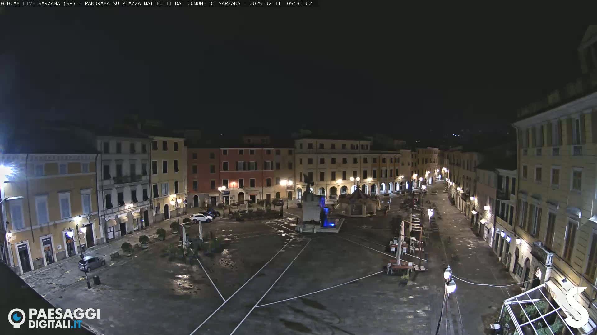 A nighttime view of a mostly empty town square with surrounding buildings lit, wet pavement, and a central fountain.