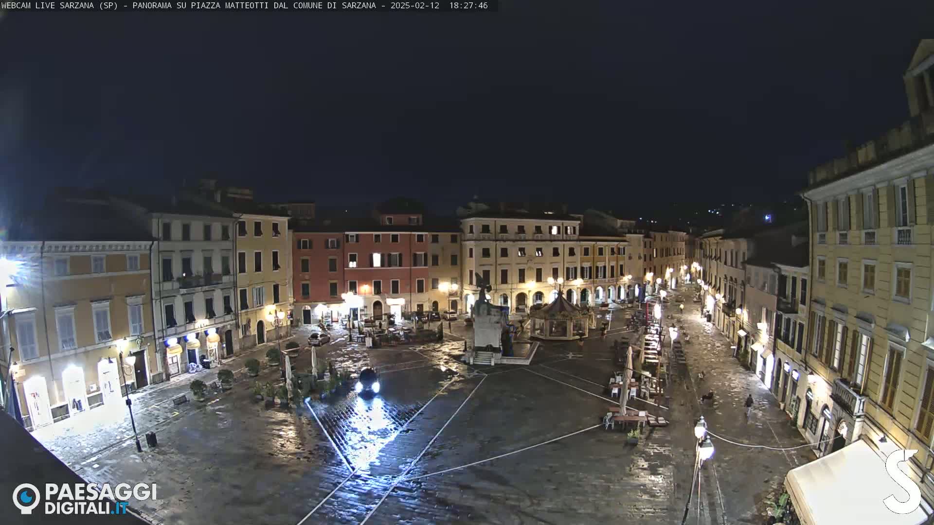 A wet, nighttime view of a town square surrounded by buildings with lights illuminating the area.