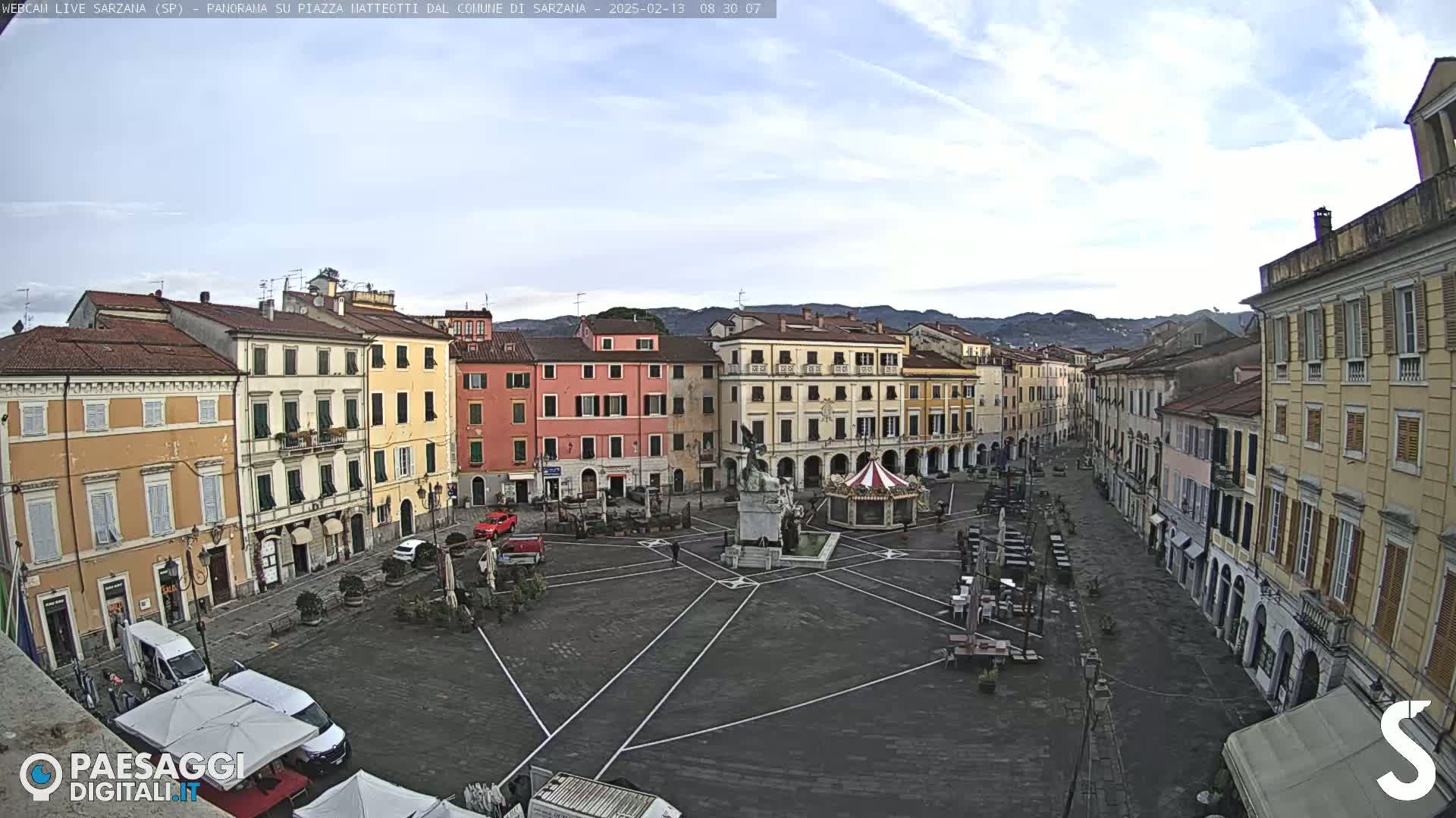 A mostly empty town square with multi-story buildings surrounding it under a partly cloudy sky.