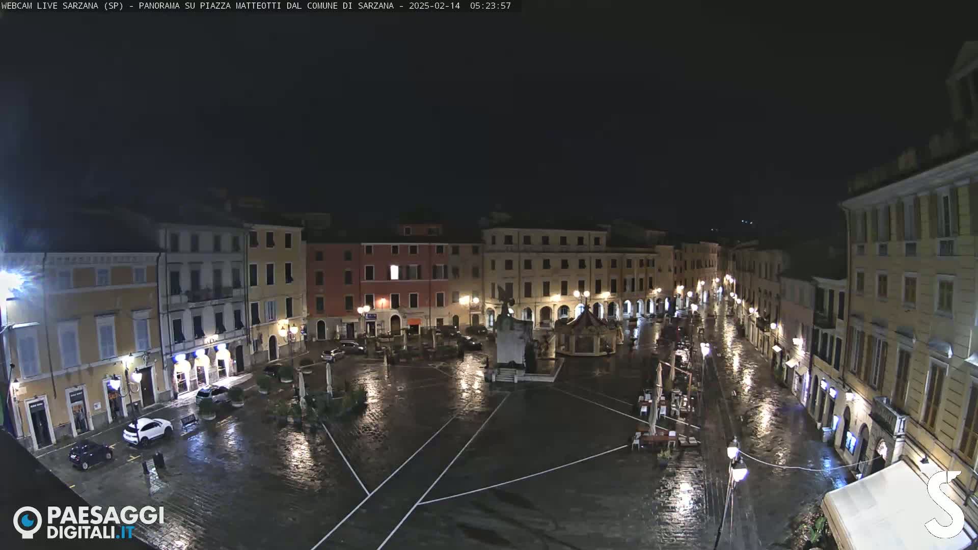 A rain-slicked town square at night, surrounded by illuminated buildings.