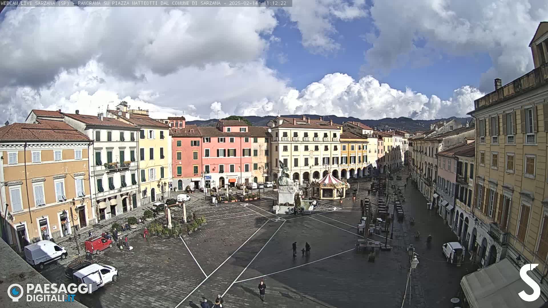 A partly cloudy day reveals a large, mostly empty town square surrounded by multi-story buildings, with a few people and vehicles visible.