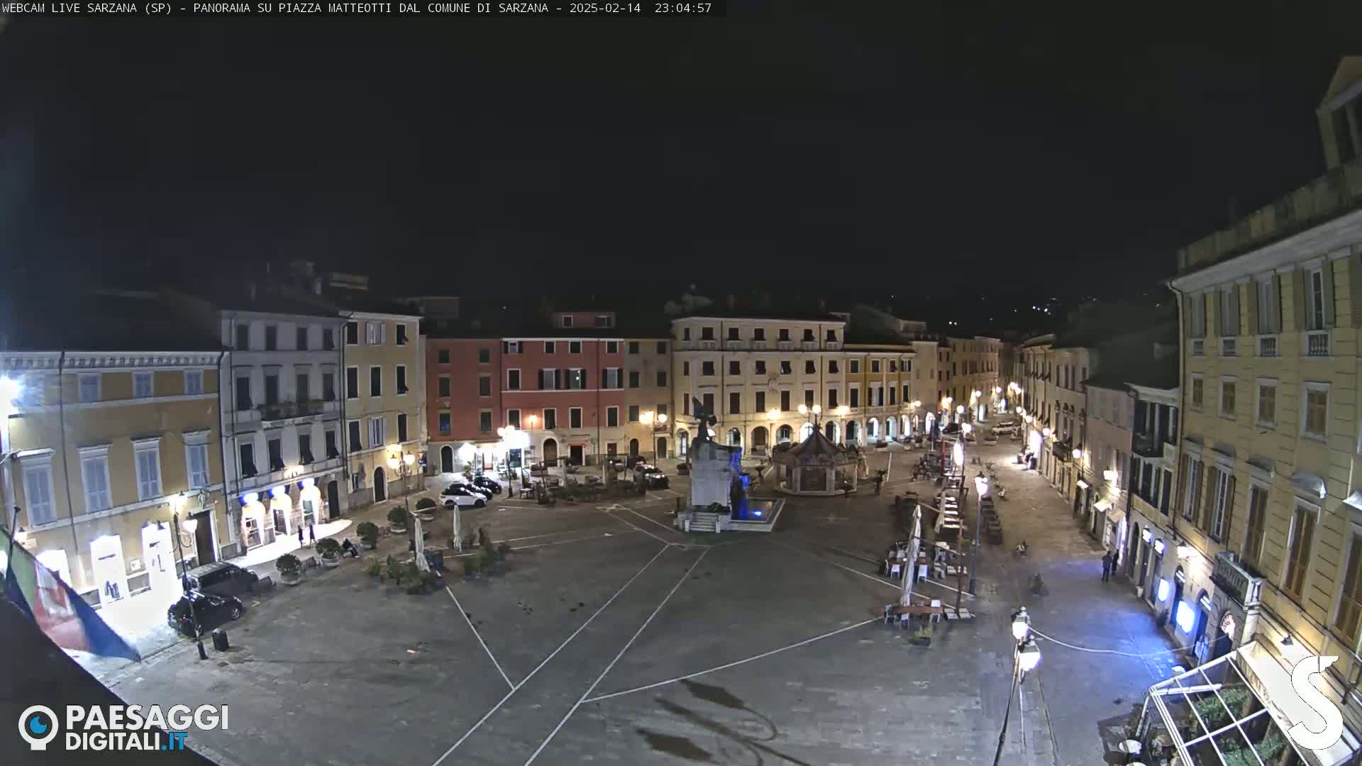 A nighttime, wide shot of a town square in Italy, featuring several buildings with illuminated windows, a central fountain, and sparse outdoor seating.