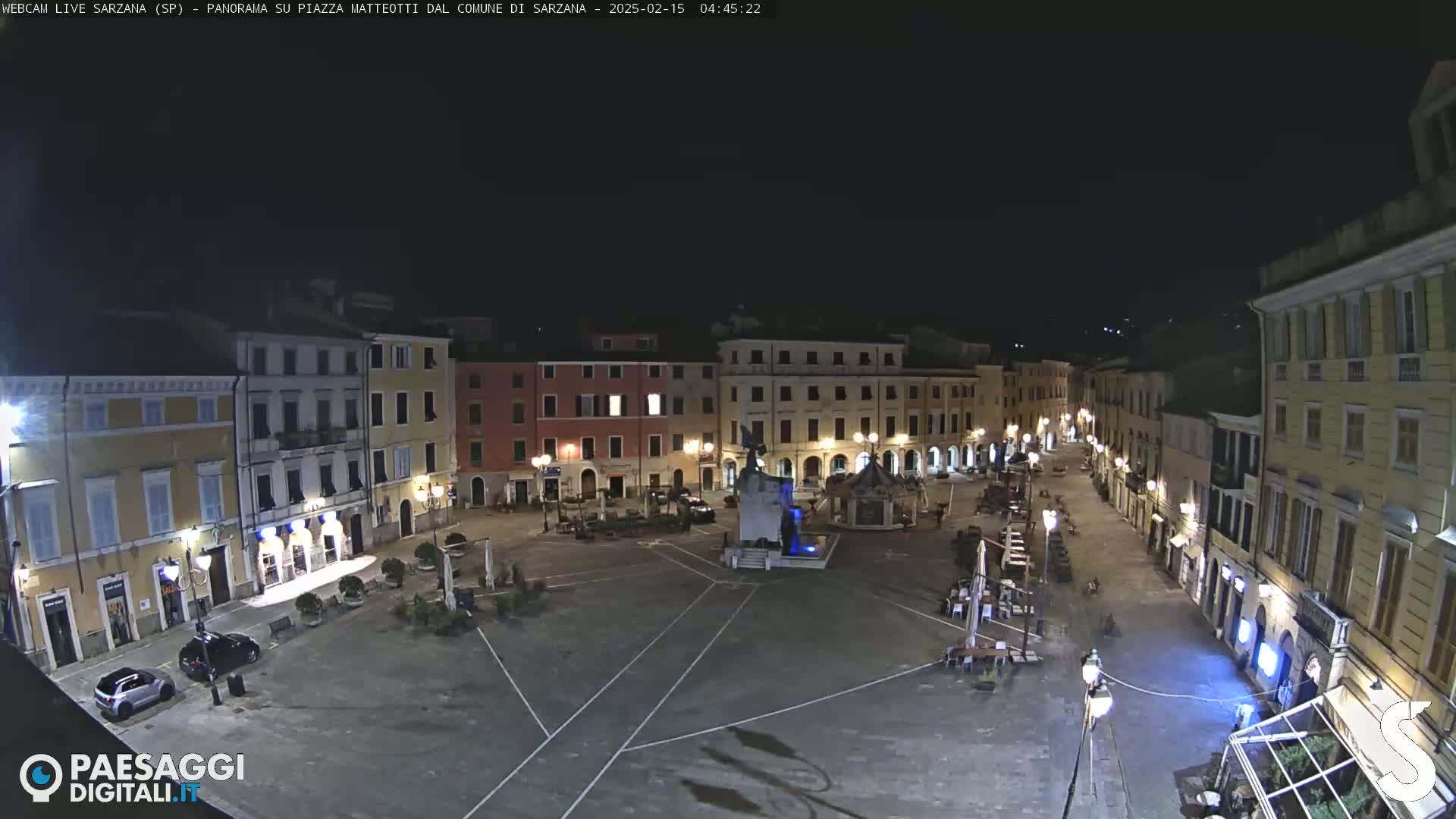 A mostly empty town square at night, illuminated by streetlights, features buildings lining its perimeter and a central fountain.