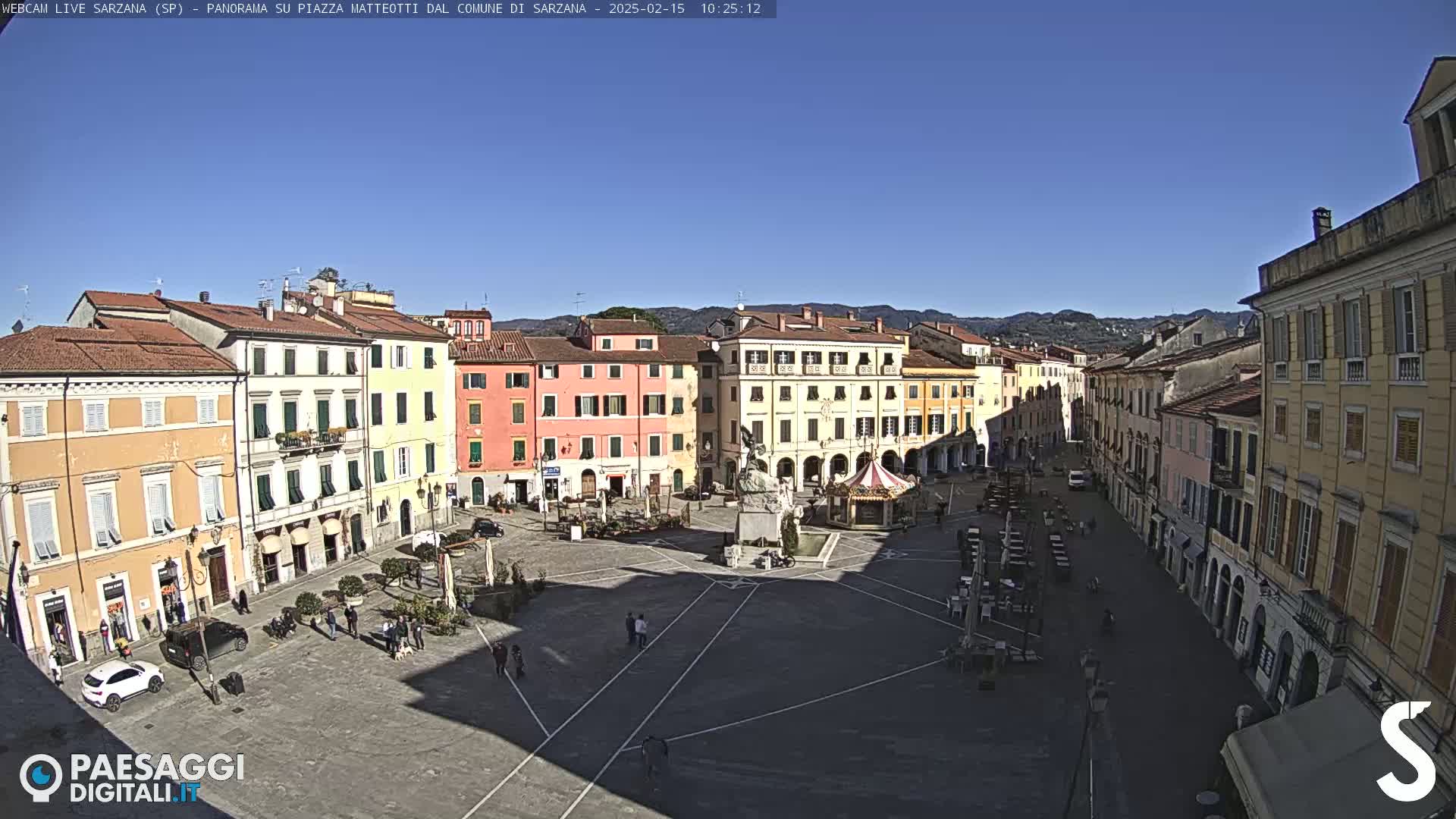 A sunny day reveals a large, mostly empty town square surrounded by multi-story buildings in a European-style town.