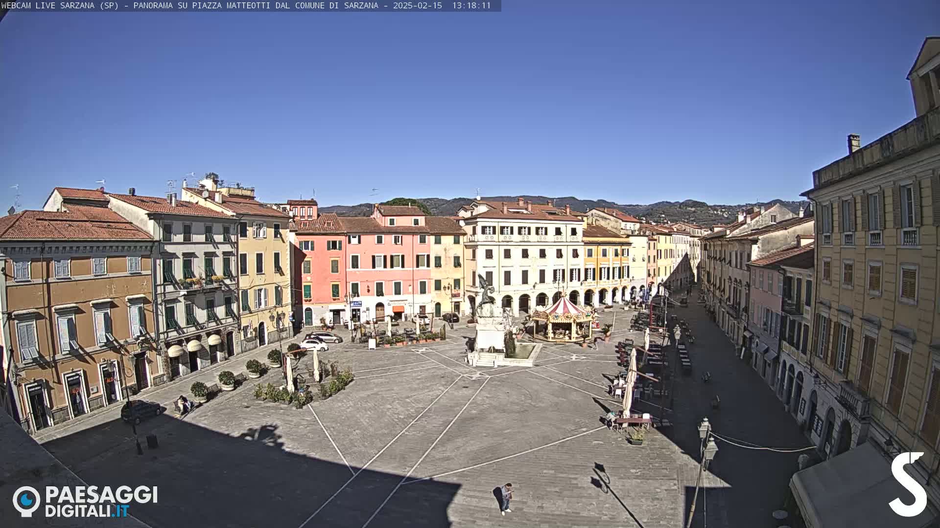 A sunny day in a town square featuring multi-story buildings, a fountain, a carousel, and several people scattered throughout the plaza.