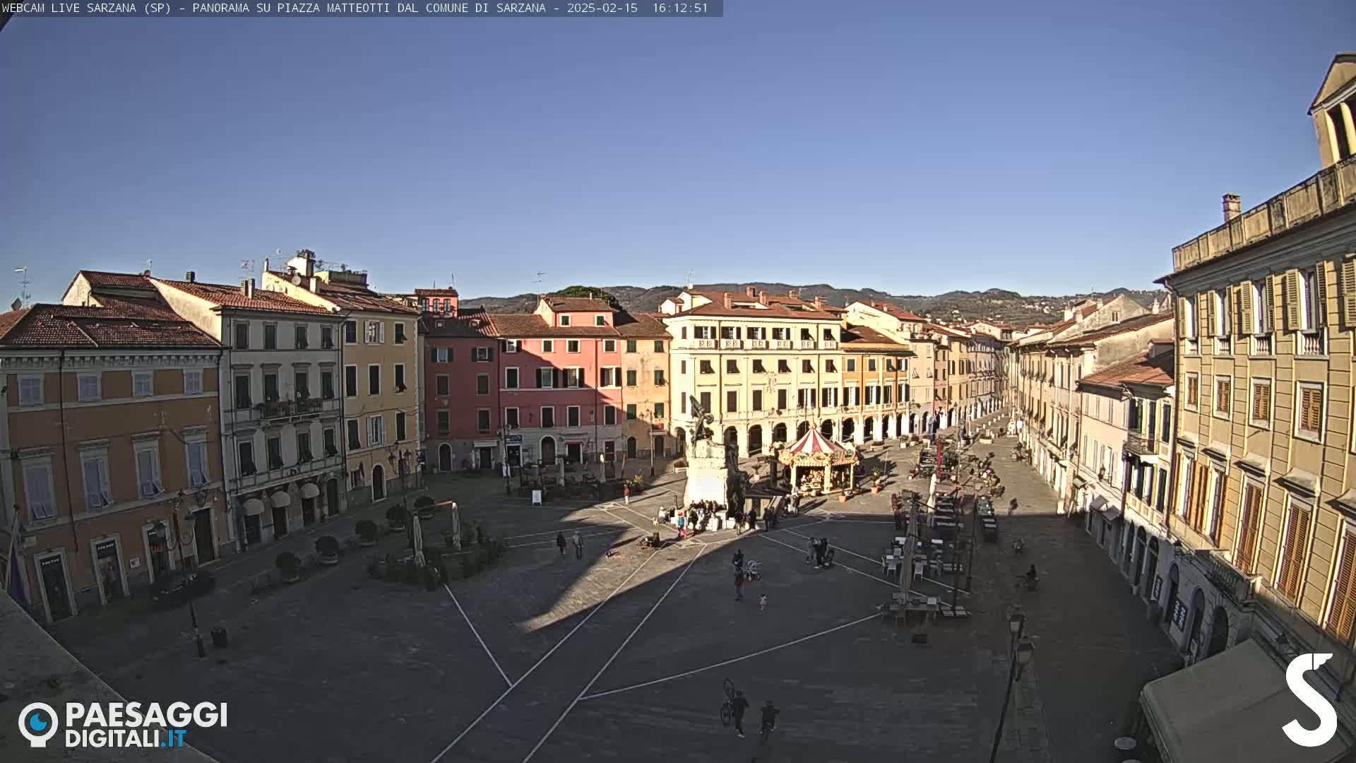 A large, paved town square is surrounded by multi-story buildings under a clear, sunny sky.