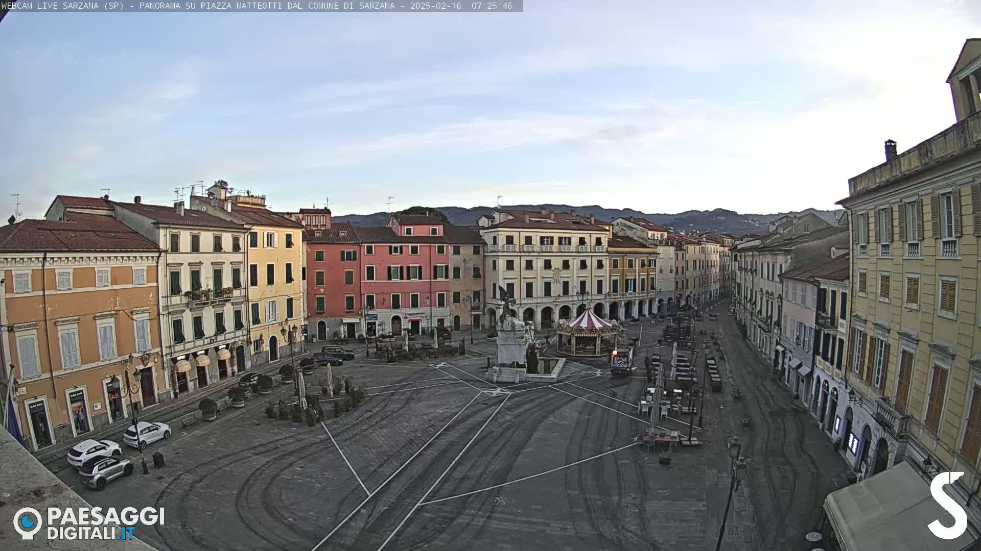 A mostly empty town square with several multi-story buildings surrounding it under a clear, dawn sky.