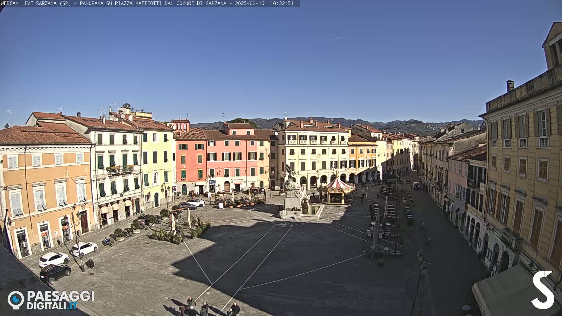 A sunny view of a large town square surrounded by pastel-colored buildings, with a few people and cars visible.