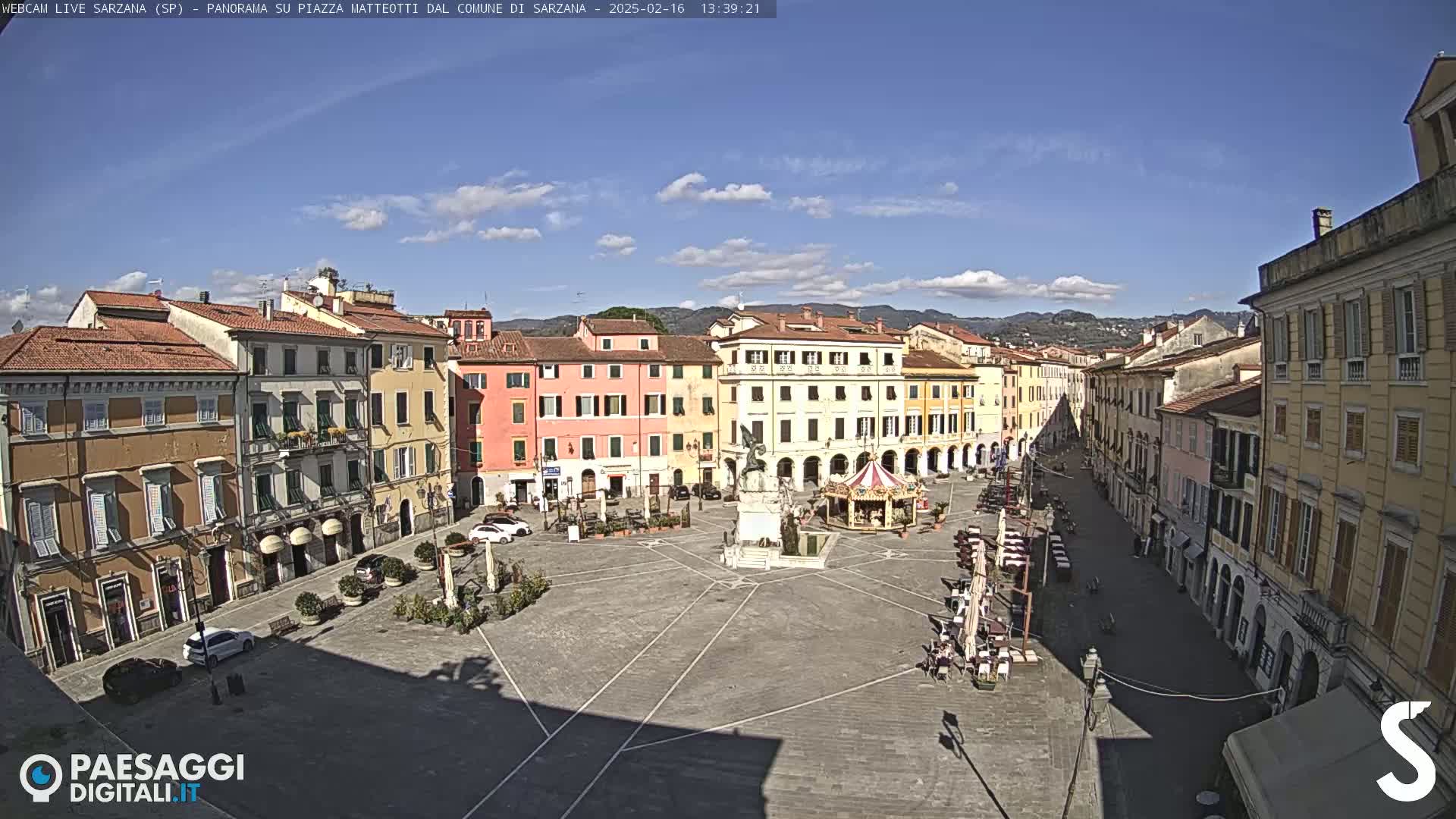 A mostly sunny day overlooks a large town square with multiple-story buildings surrounding it, a monument in the center, and a carousel.