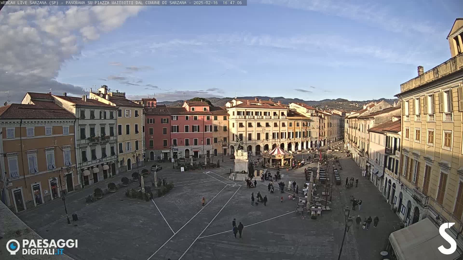 A mostly sunny day shows a large town square surrounded by multi-story buildings, with a few people walking around and some outdoor seating areas.