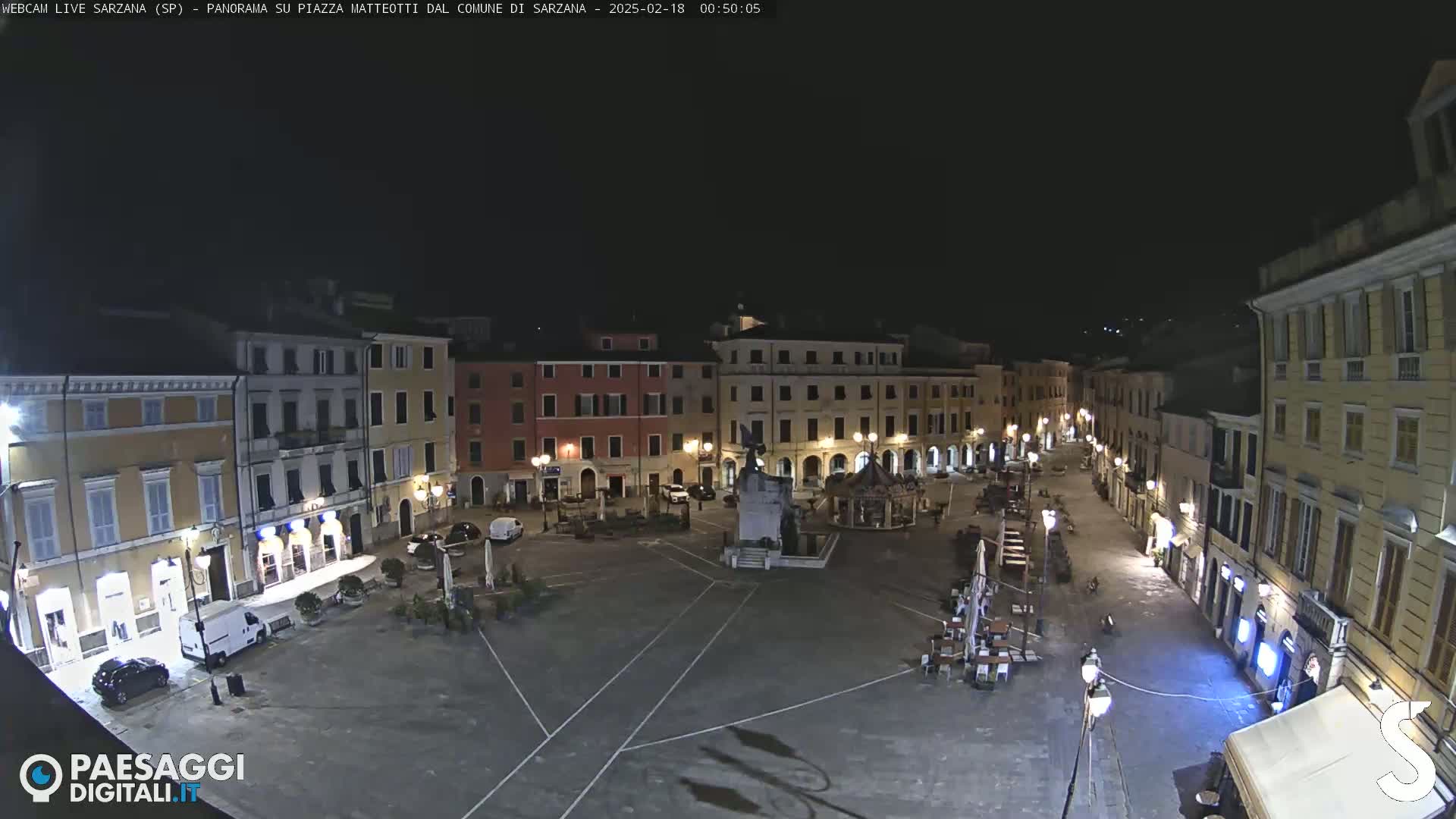 A nighttime view of a mostly empty town square surrounded by buildings, with a few cars and outdoor seating visible.