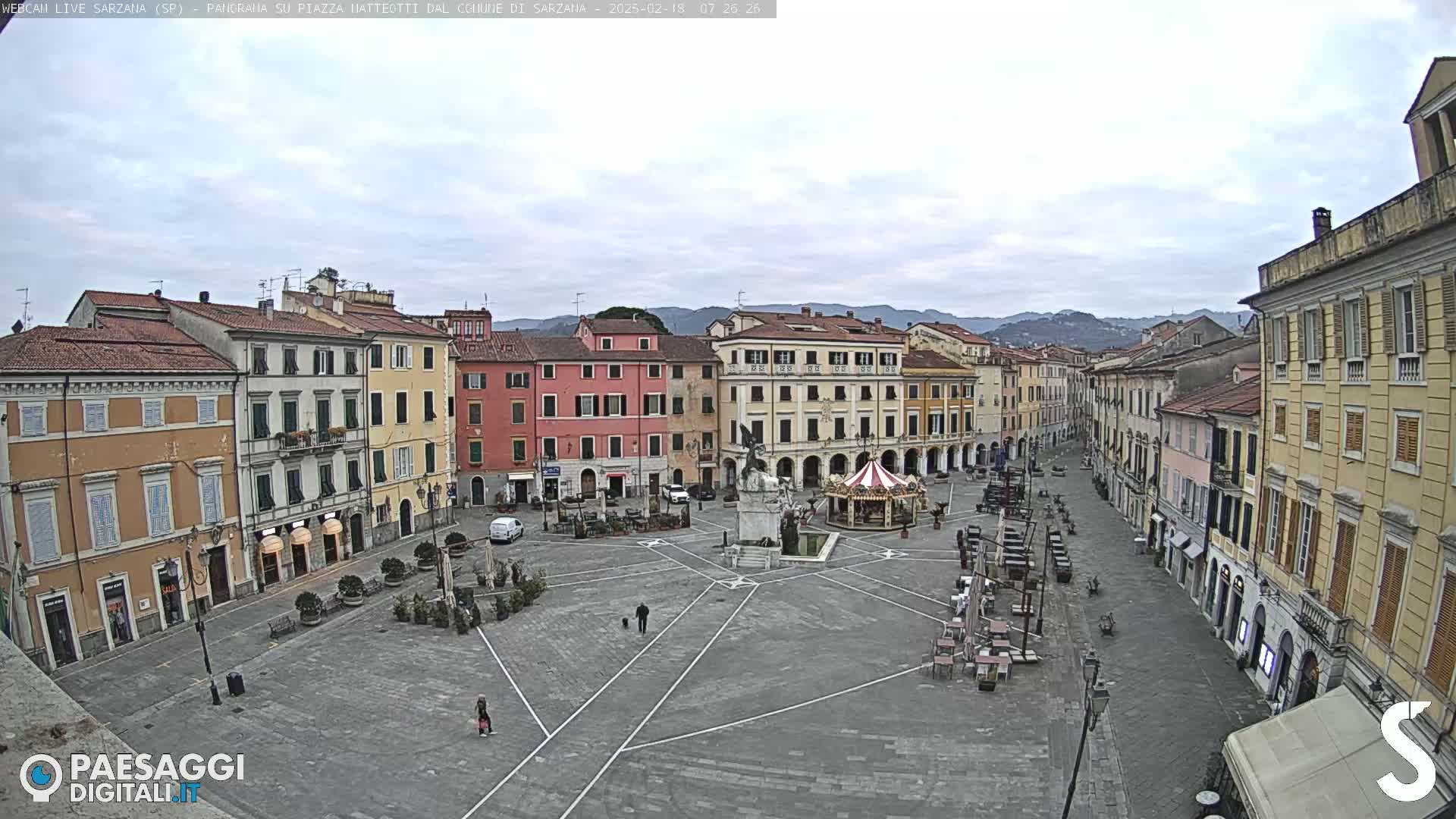 A mostly empty town square in Sarzana, Italy, is surrounded by multi-story buildings under an overcast sky.