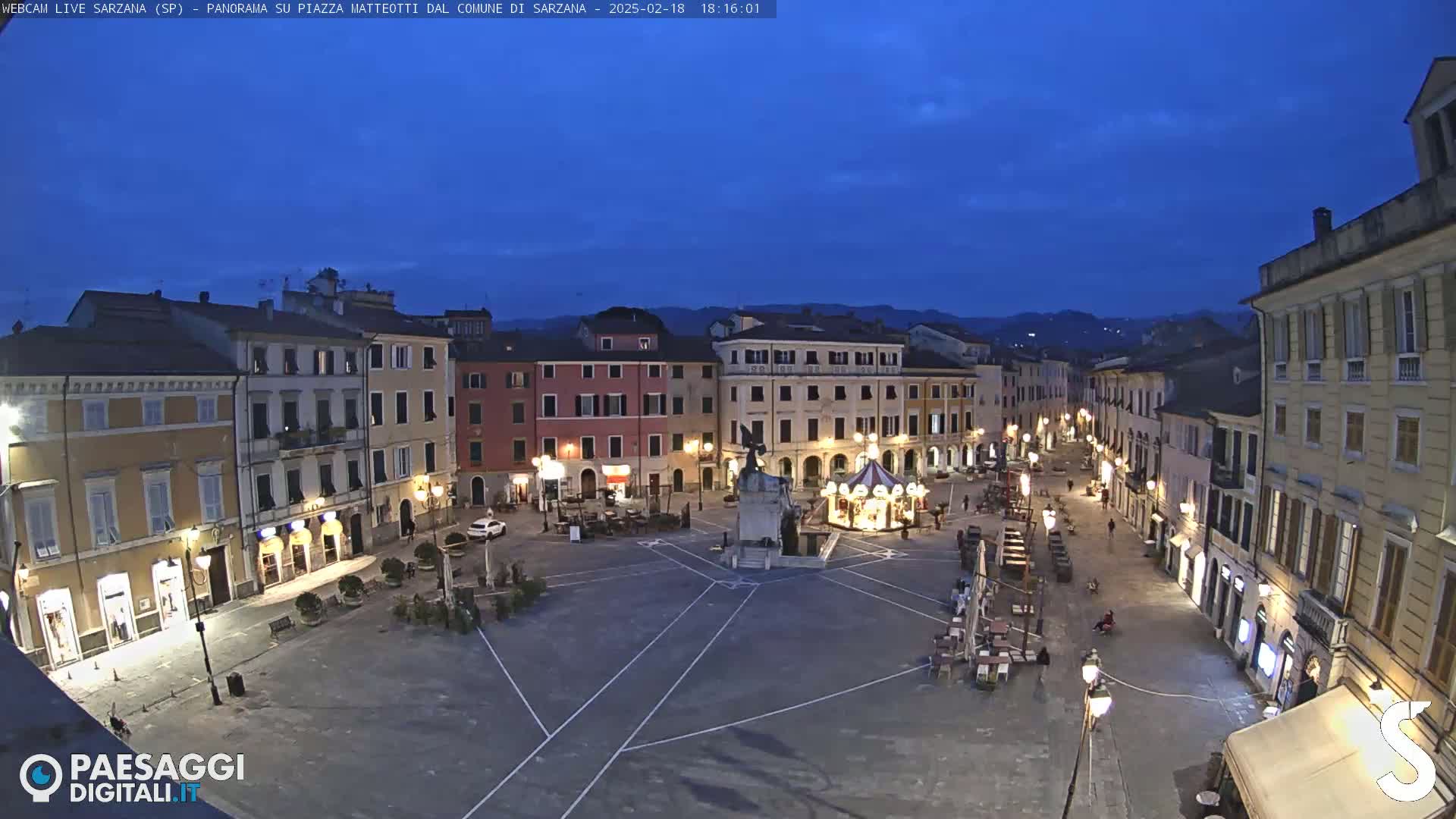 A mostly empty town square at dusk, surrounded by multi-story buildings with streetlights, features a monument and a small carousel.