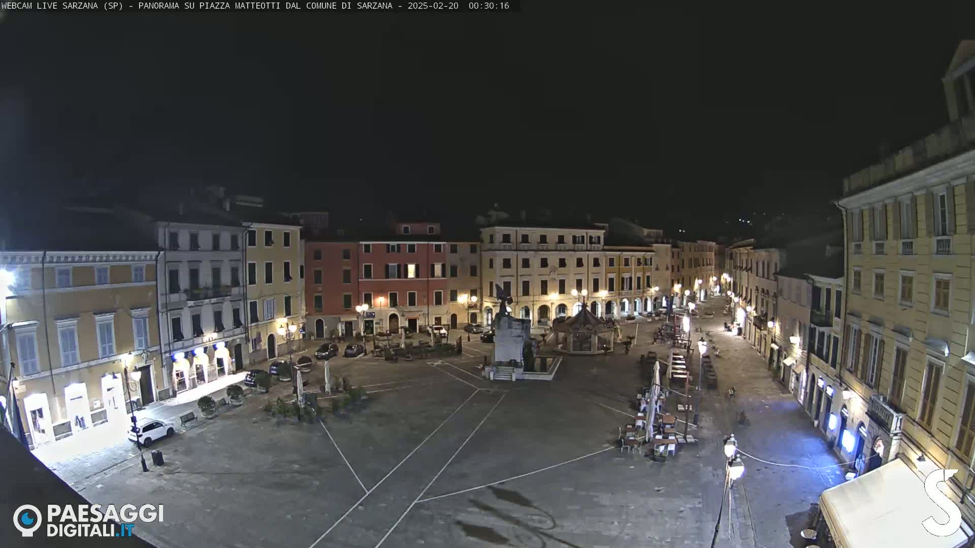 A nighttime, high-angle view of a mostly empty town square, surrounded by buildings with illuminated windows and outdoor seating areas.