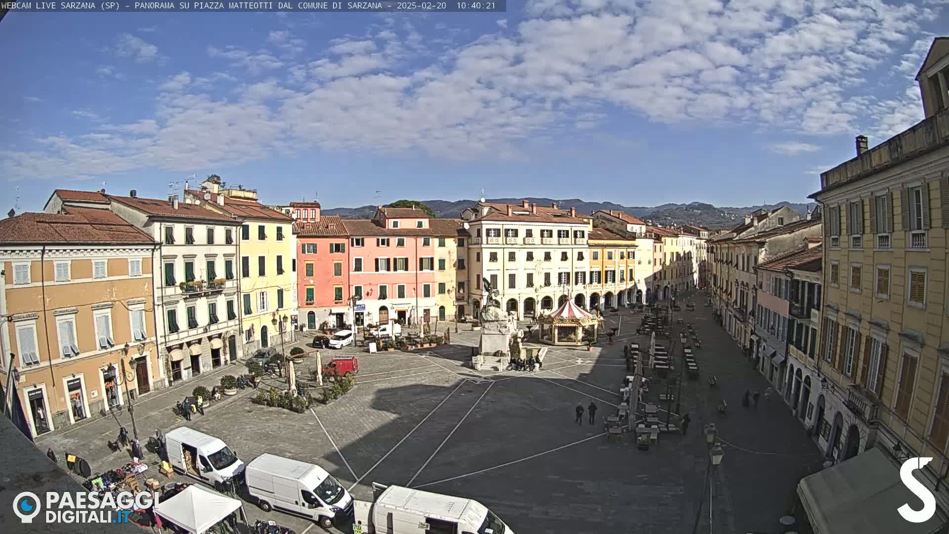 A sunny day in a large, open town square surrounded by multi-story buildings, with several vehicles parked nearby and people scattered throughout the area.