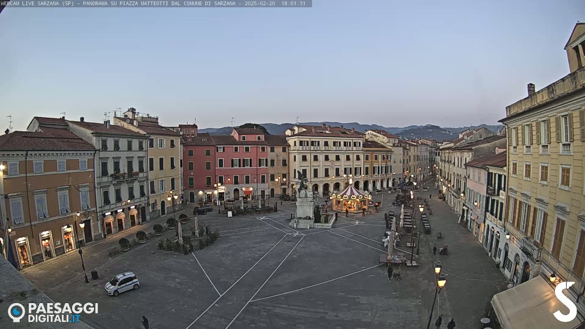 A twilight view of a town square in Italy, featuring multi-story buildings surrounding a paved plaza with a carousel and a monument, under a clear sky.