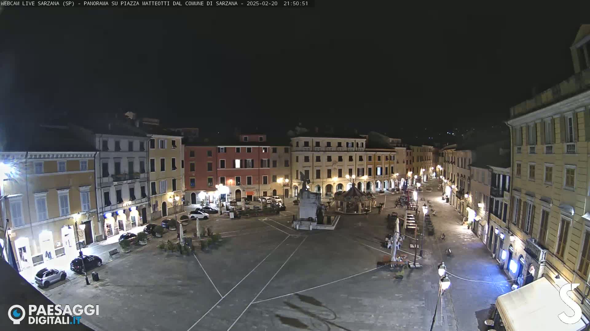 A nighttime view of a town square, illuminated by streetlights, with several buildings surrounding it and a few cars parked nearby.