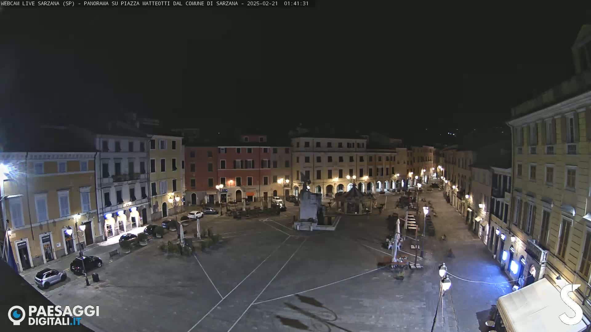 A nighttime view of a town square, illuminated by streetlights, with several buildings surrounding it and a few cars parked.