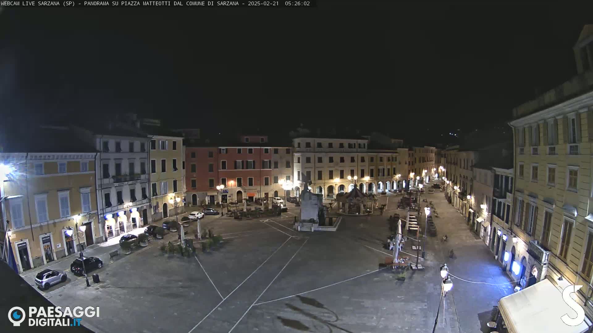 A nighttime view of a town square, surrounded by multi-story buildings, with a few cars parked and some outdoor furniture sparsely placed in the square.