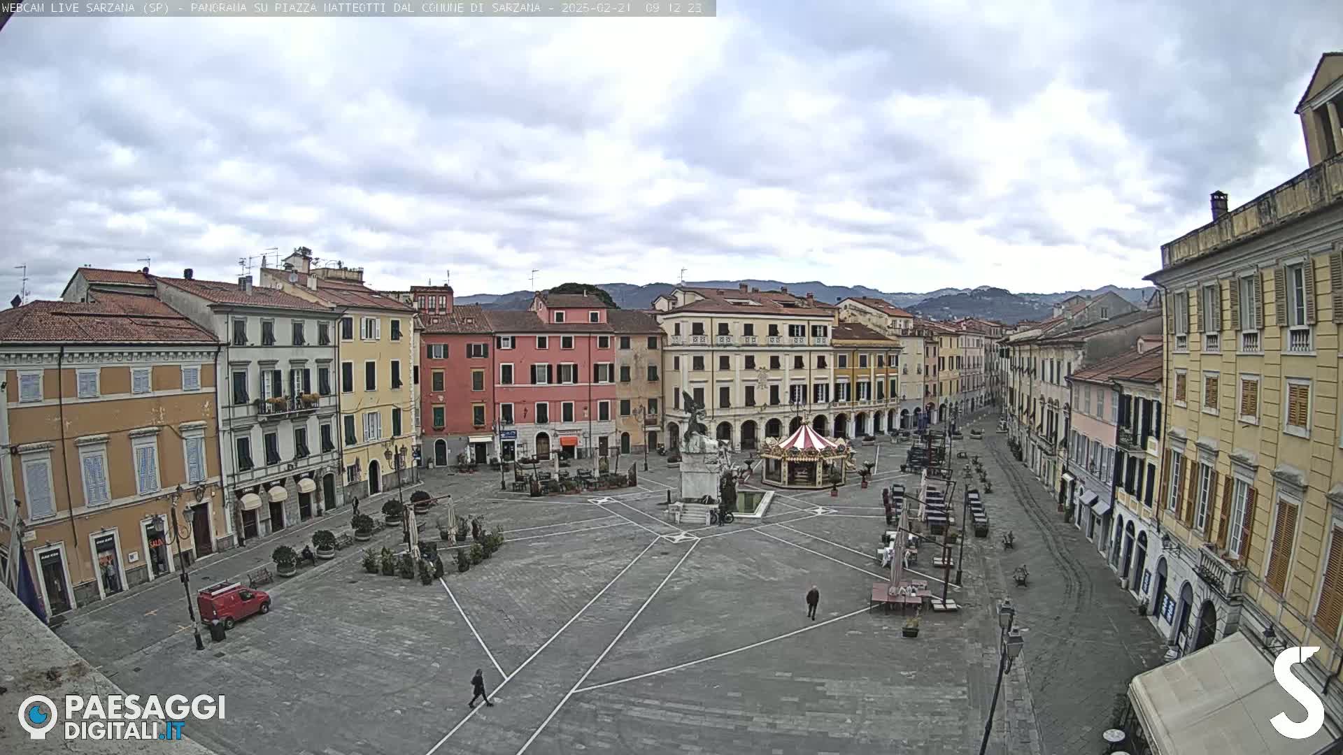 A mostly empty town square, surrounded by multi-story buildings, under a cloudy sky.