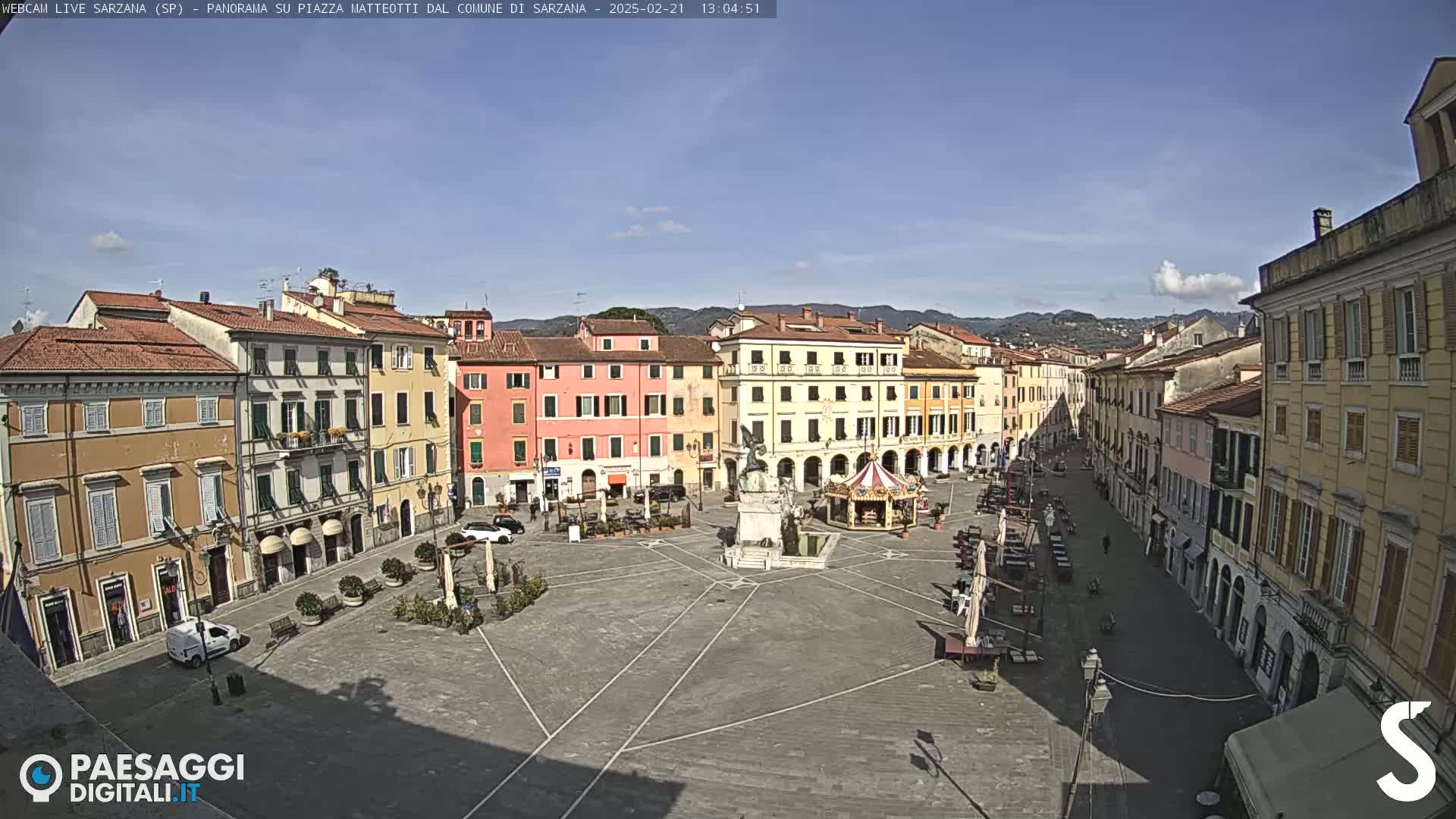 A mostly sunny day reveals a large town square surrounded by multi-story buildings, with a few cars and people visible.