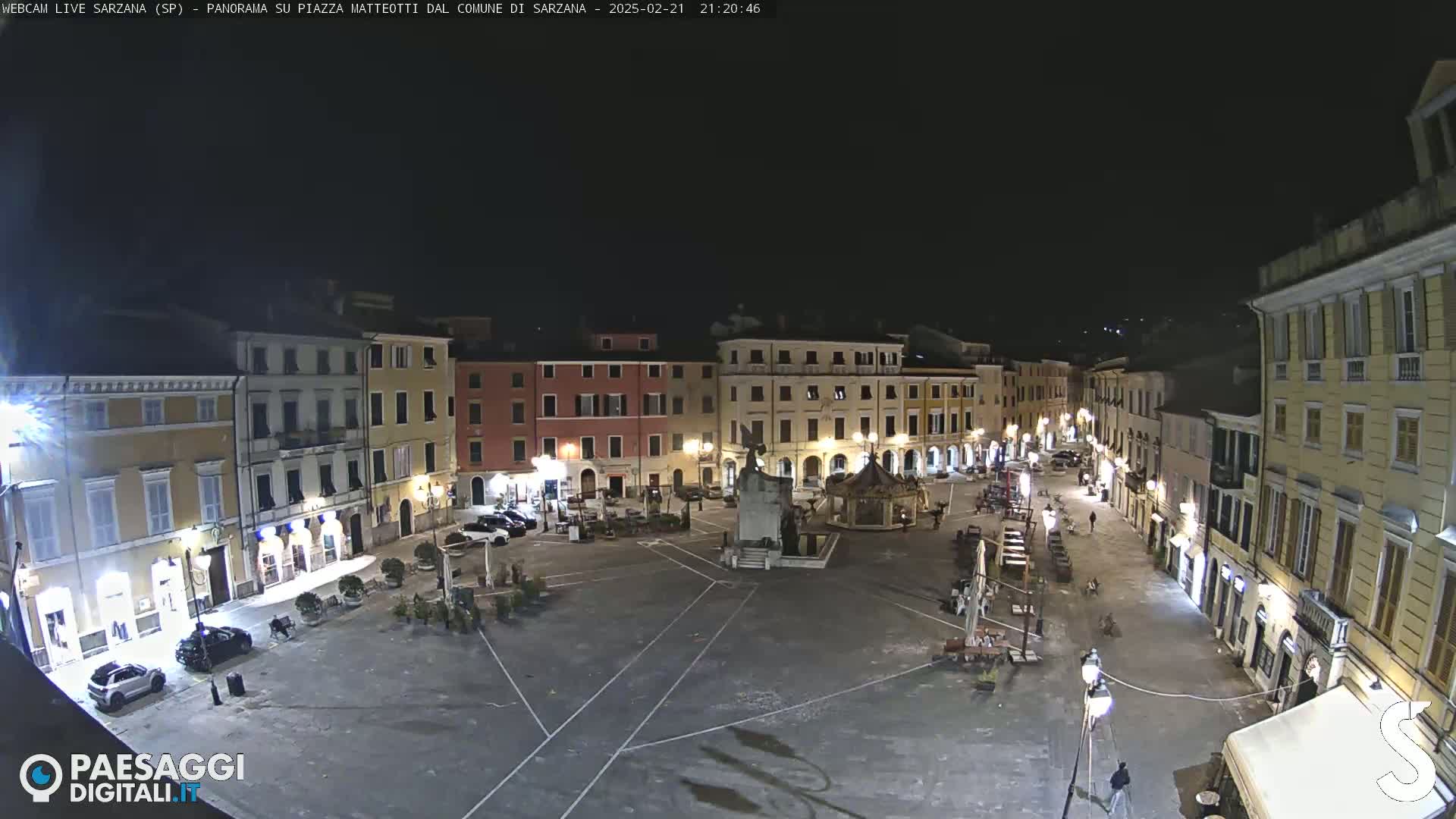 A nighttime view of a town square, illuminated by streetlights, surrounded by buildings.