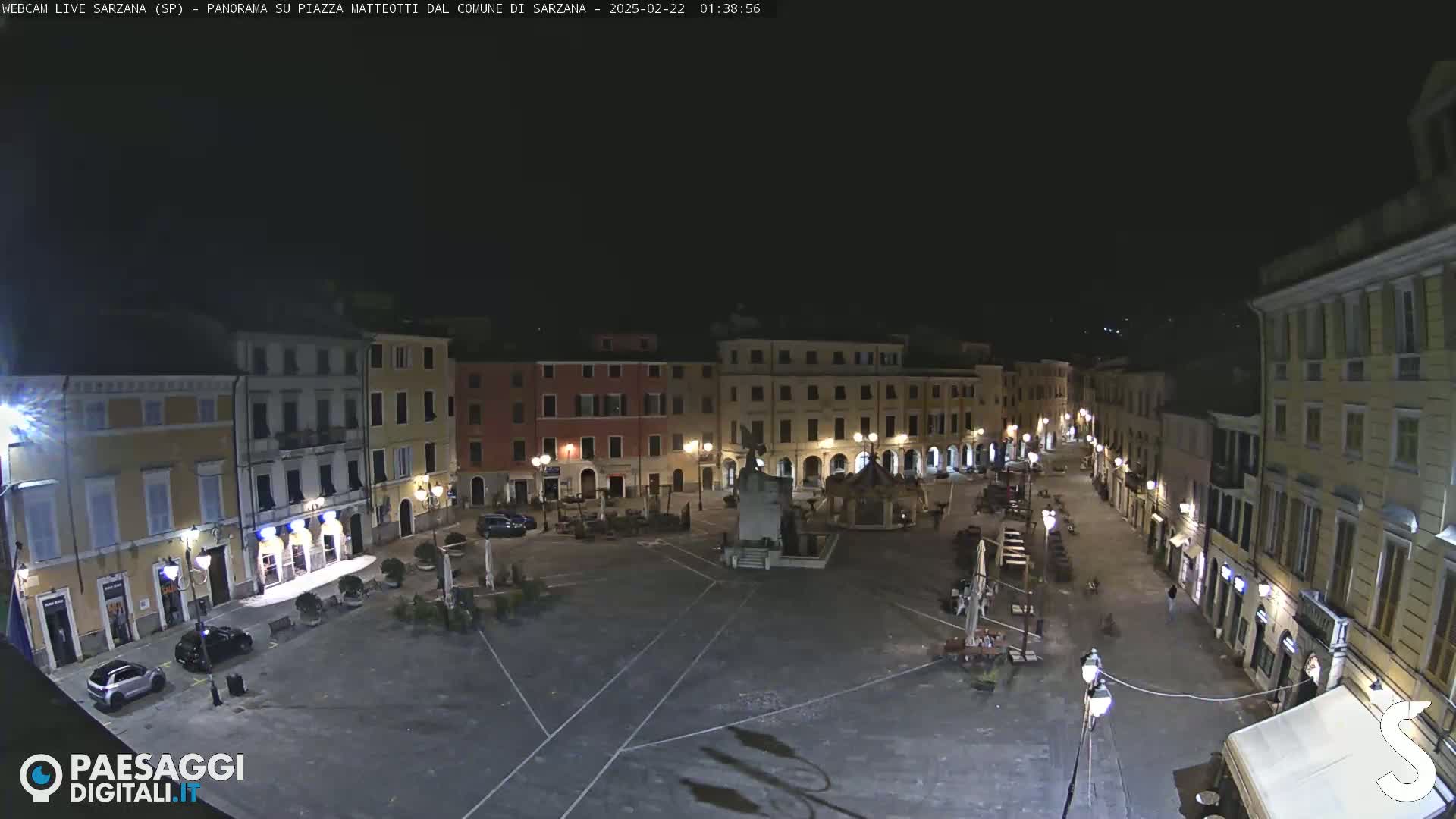 A nighttime view of a mostly empty town square, surrounded by buildings with illuminated windows and streetlights.