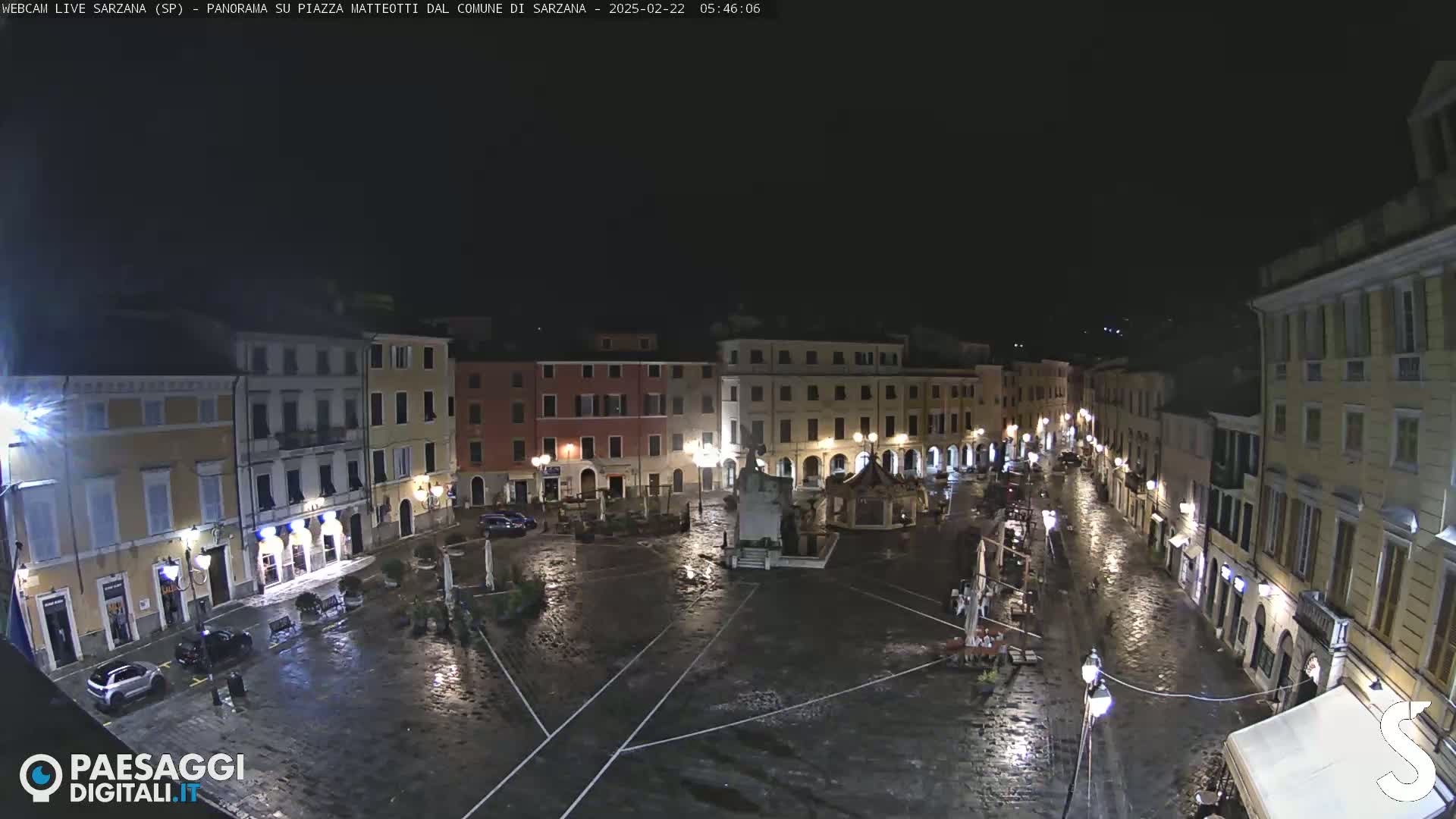 A rain-slicked town square at night, surrounded by illuminated buildings.