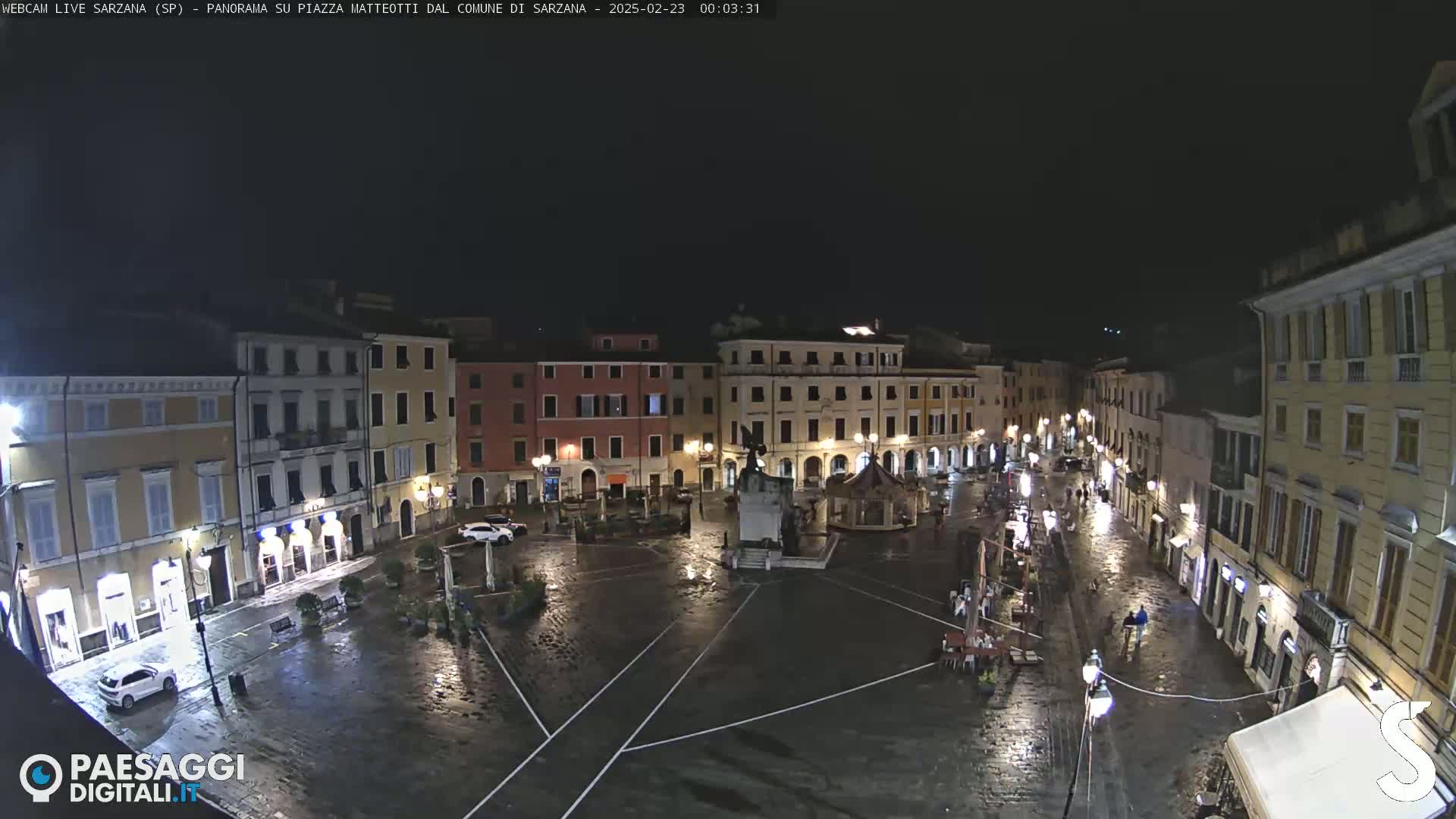 A rain-slicked town square at night, surrounded by illuminated buildings.