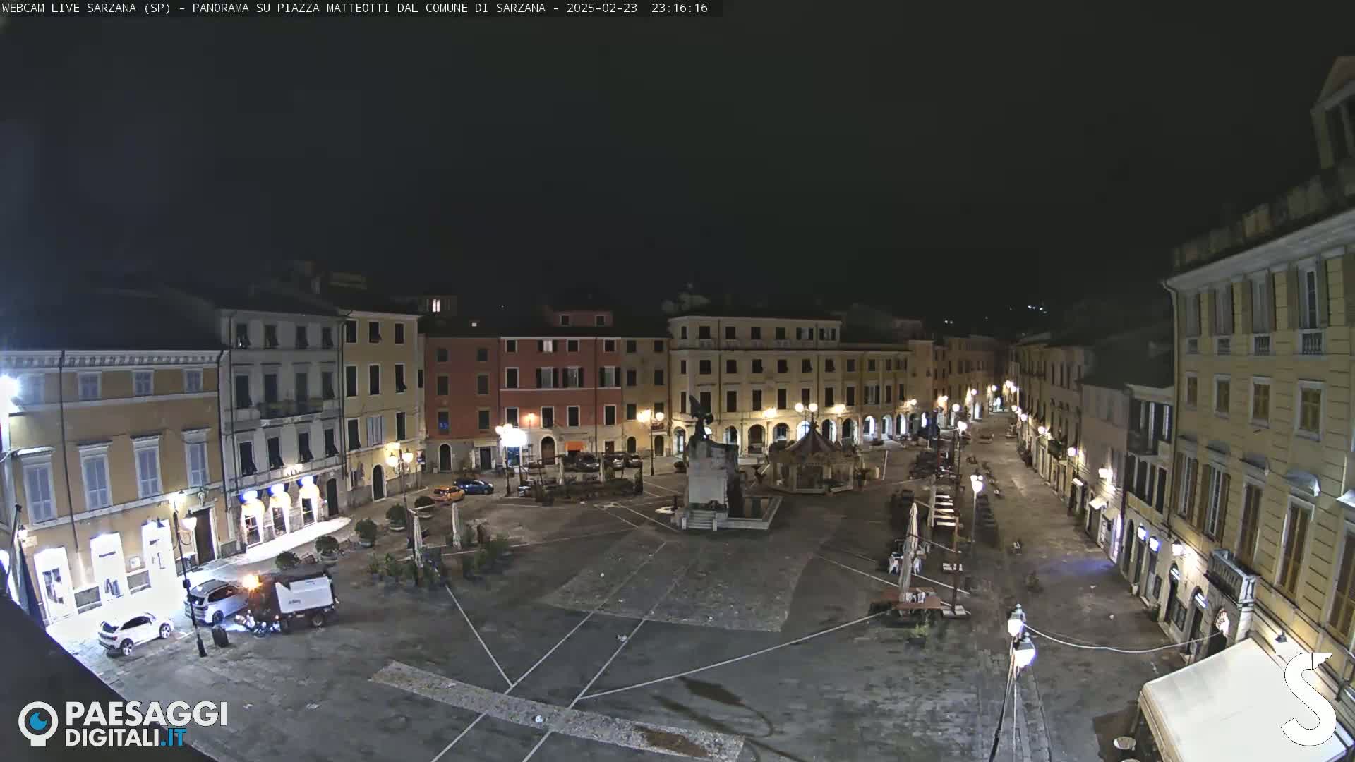 A nighttime, deserted town square, surrounded by illuminated buildings, features a central monument and a few parked vehicles.