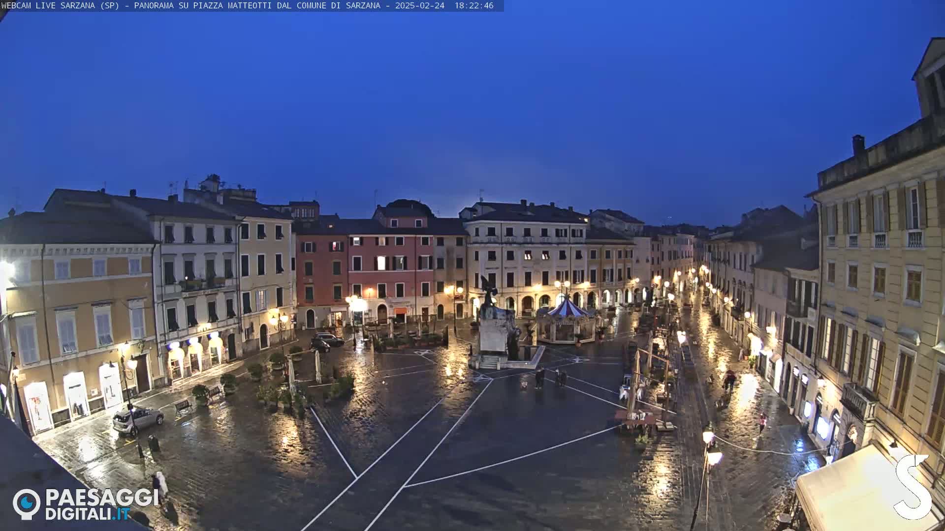 A rain-slicked town square at night, illuminated by streetlights, is surrounded by multi-story buildings.