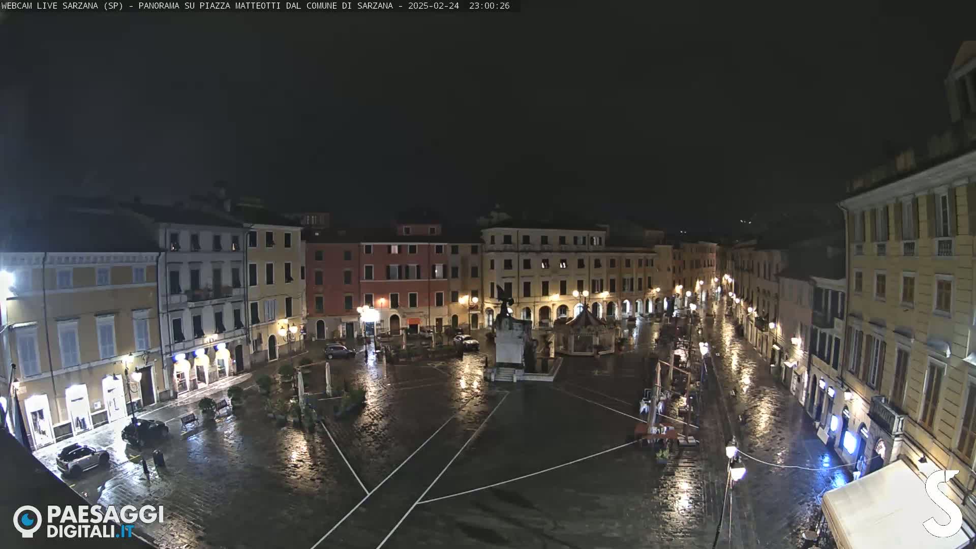 A rain-slicked town square at night, surrounded by illuminated buildings, features a central monument.