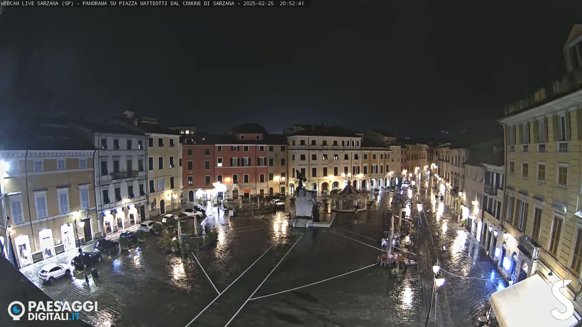 A rain-soaked town square at night, surrounded by illuminated buildings, shows cars parked along the edges and a few people walking in the wet streets.