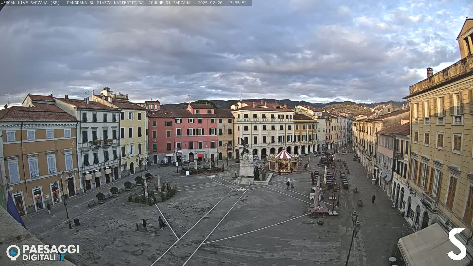 A mostly cloudy late afternoon view of a large town square in Italy, surrounded by multi-story buildings, with a monument and a small carousel in the center.